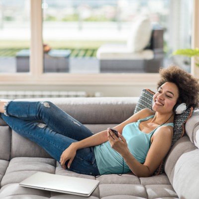 Hausratversicherung: Frau liegt auf Sofa mit Handy in der Hand und Kopfhörer auf dem Kopf